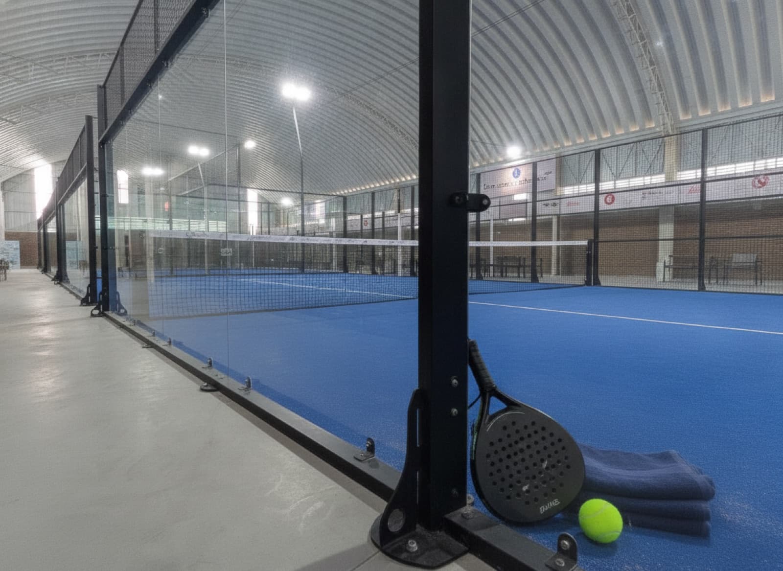 Pala y toalla recargadas en el cristal de una cancha de Padel Lomas con una pelota sobre el pasto azul