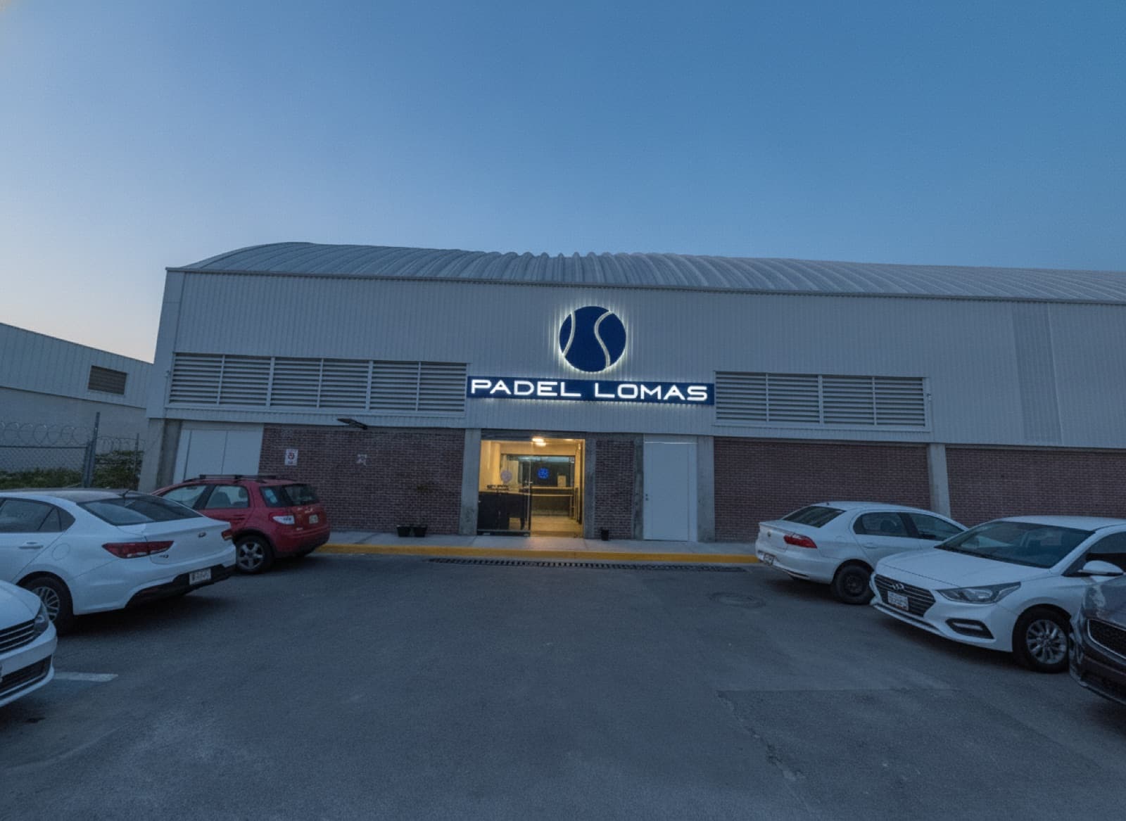 Fachada de Padel Lomas al atardecer con el letrero iluminado y autos estacionados